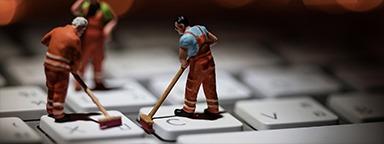 Миниатюрные люди чистят белую клавиатуру компьютера, на заднем плане мерцают оранжевые лампы