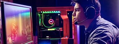 молодой геймер с гарнитурой сидит за своим столом и играет в FPS-игры; на фоне — рабочий стол его компьютера